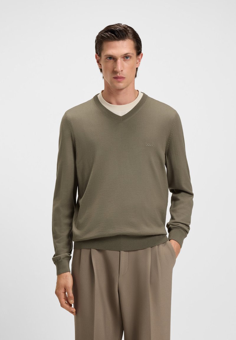Mann in olivgrünem V-Ausschnitt-Pullover über beigem Hemd mit beigen Hosen, steht mit einer Hand in der Tasche vor einem einfarbigen Hintergrund.