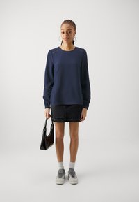 Navy blue textured sweater with long sleeves, paired with a denim skirt and gray sneakers. Holds a small black handbag.