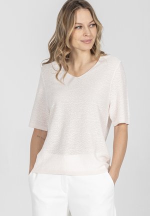 Kobieta o blond, falowanych włosach, ubrana w jasny, teksturowany krótki top z krótkim rękawem i białe spodnie, stoi z rękami w kieszeniach, patrząc w prawo.