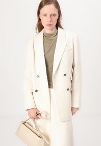Dvojradový biely blazer z textúrovanej látky, v kombinácii s tlmeným zeleným topom. Modelka drží béžovú obdĺžnikovú kabelku. Jednoduché pozadie.