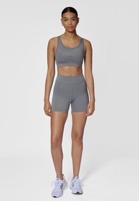 Brassière de sport grise avec texte "HUMMEL", short taille haute assorti gris. Tissu lisse et extensible ; coupe ajustée ; portée avec des chaussures de sport légères.