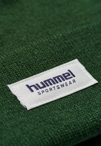 Grön textil med en strukturerad yta som har en vit etikett med texten "hummel SPORTSWEAR" i blått, sytt vid kanten.