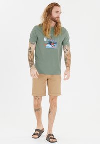 Vihreä puuvillapaita, jossa on graafinen tuuletuskuva, yhdistetty khaki-shortseihin. Ruskeat sandaalit, joissa on solk yksityiskohta.