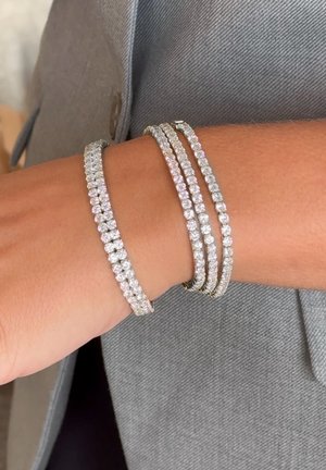 Tre silverfärgade armband med klara kristalldetaljer, med en delikat, texturerad design. Bärs på ett handled mot en grå tygbakgrund.