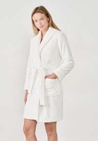 Accappatoio bianco in pile con colletto a scialle, cintura da annodare e tasche frontali. Texture morbida, design fino al ginocchio e dettagli a coste sui polsini.