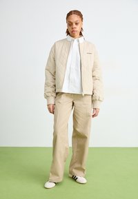 Giubbotto bomber beige trapuntato con polsini a coste, abbinato a una camicia bianca a righe e pantaloni beige. Il modello indossa scarpe bianche con dettagli neri.