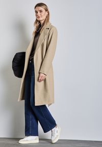 Beżowy wełniany płaszcz o dopasowanym kroju, długich rękawach i klapach. W zestawie z ciemnoniebieskimi, szerokimi dżinsami i białymi trampkami.