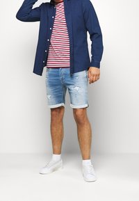 Mężczyzna w niebieskiej koszuli dżinsowej noszący ją na czerwono-białej koszulce w paski, podartych jasnoniebieskich szortach dżinsowych, białych skarpetkach i białych trampkach.