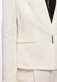 Blazer de cor creme com tecido suave, colarinho arredondado, um bolso frontal, e detalhe de fecho em tom prateado.
