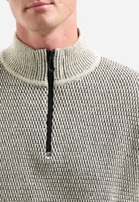 Man in een beige en zwart gestructureerde gebreide trui met een geribde kraag en een zwarte rits tot de helft.