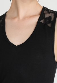 Svart ärmlös topp med V-ringning och spetsdetaljer på axlarna, bärs av en person som visar hals och övre bröstområde.