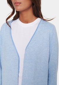 Ljusblå cardigan med v-ringning, tillverkad av mjukt material. Lagd över en vit t-shirt med rund halsringning. Slät textur, minimal detaljering.