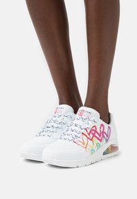 Weiße Sneakers mit bunten, abstrakten Designs, strukturiertem Material, Schnürverschluss und einer sichtbaren Luftpolsterung in der Ferse.