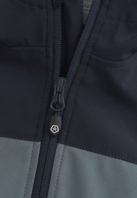 Mörkblå och grå blandad tygjacka med dragkedja och texturerad dragflik. Har platta sömmar och ficka ovanför dragkedjan. Inga mönster synliga.