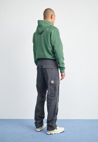 Sweatshirt à capuche verte associé à un pantalon cargo gris avec poches et des baskets blanches avec des accents jaunes. Tissu lisse et coupe structurée.