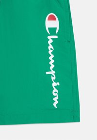 Zelené šortky z ľahkého materiálu s bielym kurzívnym logom "Champion" a emblémom s červeným akcentom. Hladká textura, voľný strih.