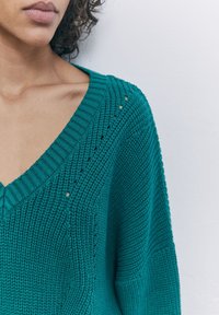 Teal neulepusero, jossa on syvä V-pääntie, teksturoitu ribbikuviointi ja pieniä avoimia yksityiskohtia hartioilla. Pehmeä, kevyt materiaali.