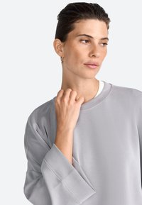 Sudadera gris claro con cuello redondo, corte relajado y mangas anchas. Textura suave con detalles de costura en los puños.