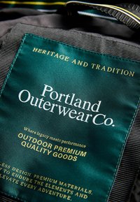 Zelená textilní etiketa s bílým a zlatým textem. Text obsahuje "Portland Outerwear Co." a fráze o outdoorovém výkonu a kvalitních výrobcích.