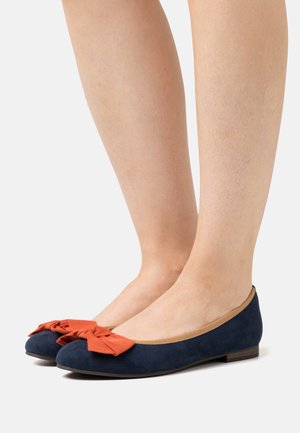 Pieds portant des ballerines en daim bleu marine avec des nœuds en tissu orange et des bordures beiges sur fond blanc.