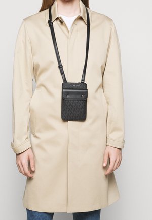 Person trägt einen beigen Trenchcoat und blaue Jeans mit schwarzer Michael Kors Crossbody-Handytasche auf Brusthöhe.