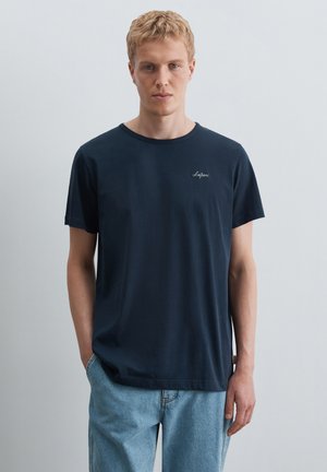 Ung mand med kort blondt hår iført en ensfarvet mørkeblå T-shirt og lyseblå jeans, stående mod en ensfarvet baggrund.