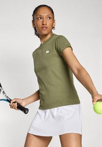 Grønn bomulls t-skjorte med korte ermer og en liten hvit logo, sammen med et hvitt sportsskjørt. Holder en tenniskølle og ball.