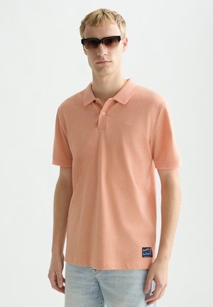 Jeune homme portant des lunettes de soleil, un polo manches courtes couleur pêche et un jean bleu clair, debout devant un fond blanc uni.