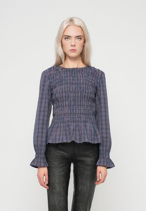 Blouse à carreaux bleu marine et rose avec design smocké, encolure ronde et manches évasées, associée à un pantalon texturé sombre.