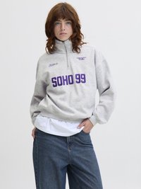 Mladý člověk s červenými vlasy, oblečený v světle šedém mikině s krátkým zipem, na které je nápis "SOHO 99" v purpurové barvě, volných džínách a bílém tričku pod tím.