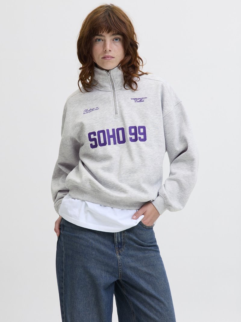 Mladý člověk s červenými vlasy, oblečený v světle šedém mikině s krátkým zipem, na které je nápis "SOHO 99" v purpurové barvě, volných džínách a bílém tričku pod tím.