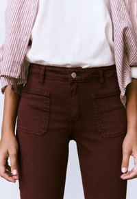 Persona que lleva pantalones de tiro alto de color rojo oscuro con bolsillos delanteros y una camisa blanca debajo de una chaqueta a rayas rojas y blancas.