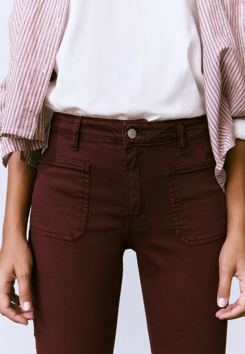 Persona que lleva pantalones de tiro alto de color rojo oscuro con bolsillos delanteros y una camisa blanca debajo de una chaqueta a rayas rojas y blancas.