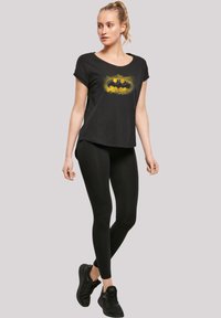Svart t-skjorte med kort erme og en gul Batman-logo, kombinert med svarte leggings og svarte joggesko. Glatt stoff og avslappet passform.