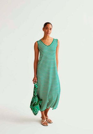 Mujer con vestido sin mangas de rayas verdes y blancas sosteniendo un bolso con estampado verde, de pie frente a un fondo liso.