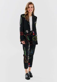 Czarny garnitur z kolorowym kwiatowym haftem, składający się z marynarki i tapered spodni. Materiał ma gładką teksturę, zestawiony z błyszczącymi czarnymi mokasynami.