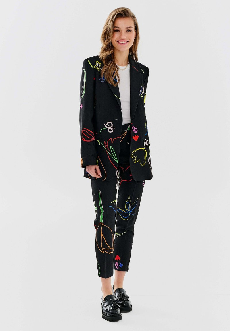 Czarny garnitur z kolorowym kwiatowym haftem, składający się z marynarki i tapered spodni. Materiał ma gładką teksturę, zestawiony z błyszczącymi czarnymi mokasynami.