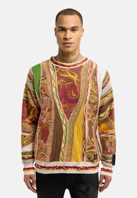 Pull en tricot coloré avec des motifs complexes en rouge, jaune et vert. Caractéristiques de tissu texturé, col rond et finitions contrastées.