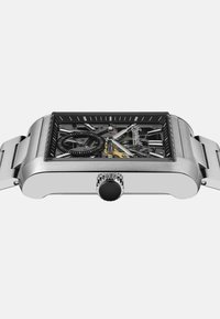 Srebrny prostokątny zegarek z przezroczystym szkieletowym cyferblatem, czarnymi akcentami i teksturowaną koroną. Bransoleta ze stali nierdzewnej dopełnia całość.