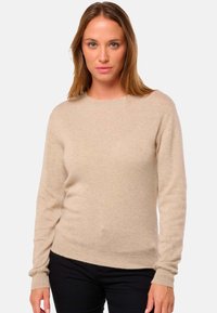 Pull en tricot beige à manches longues, décolleté rond et poignets côtelés. Texture douce avec une coupe ajustée, associé à un pantalon noir.