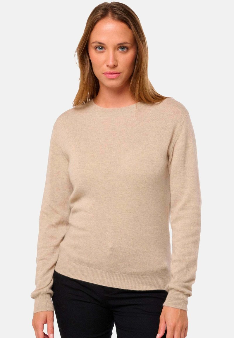 Pull en tricot beige à manches longues, décolleté rond et poignets côtelés. Texture douce avec une coupe ajustée, associé à un pantalon noir.