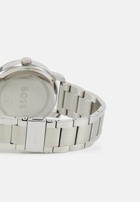 Reloj de acero inoxidable plateado con una correa suave y caja redonda, que presenta un logo en la parte posterior y un diseño minimalista. Resistente al agua.