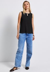 Musta hihaton paita, jossa on avainaukko-aukko, yhdistettynä vaaleansinihin leveälahkeisiin farkkuihin ja mustiin litteisiin sandaaleihin. Sujuva kankaan rakenne.