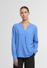 Moteris stovi su viena ranka kišenėje, vilkinti ilgomis rankovėmis šviesiai mėlyną bliusoną ir juodas kelnes prieš paprastą šviesią fono.