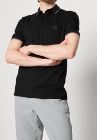Homme les bras croisés portant un polo noir à manches courtes avec bordure grise et pantalon gris clair sur un fond uni.