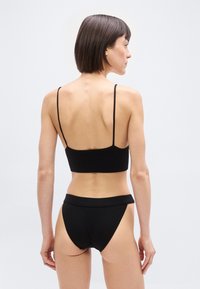 Γυναίκα με κοντά καστανά μαλλιά που φοράει μαύρο ριπ μαγιό δύο κομματιών, εμφανιζόμενη από πίσω πάνω σε απλό φωτεινό φόντο.