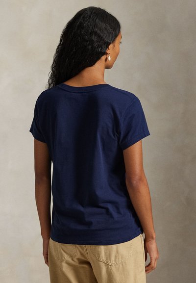 Γυναίκα με μακριά σγουρά μαλλιά, που φοράει μπλε κοντομάνικο μπλουζάκι και μπεζ παντελόνι, στέκεται με την πλάτη προς την κάμερα.