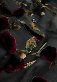 Schwarzer transparenter Stoff mit botanischer Bestickung in tiefrot und Gold, versehen mit Knöpfen und einer zarten Textur. Rosen- und Blattmuster sind sichtbar.