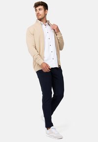 Beige gebreide cardigan met geribde manchetten en kraag, gedragen over een witte button-up shirt met donkere knopen, gecombineerd met marineblauwe broek en lichte sneakers.
