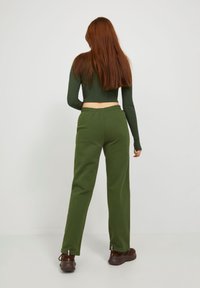 Frau mit langen roten Haaren, die ein dunkelgrünes langärmliges Crop-Top, olivgrüne Hosen und braune Schuhe trägt, steht mit dem Gesicht weggewandt vor einem weißen Hintergrund.